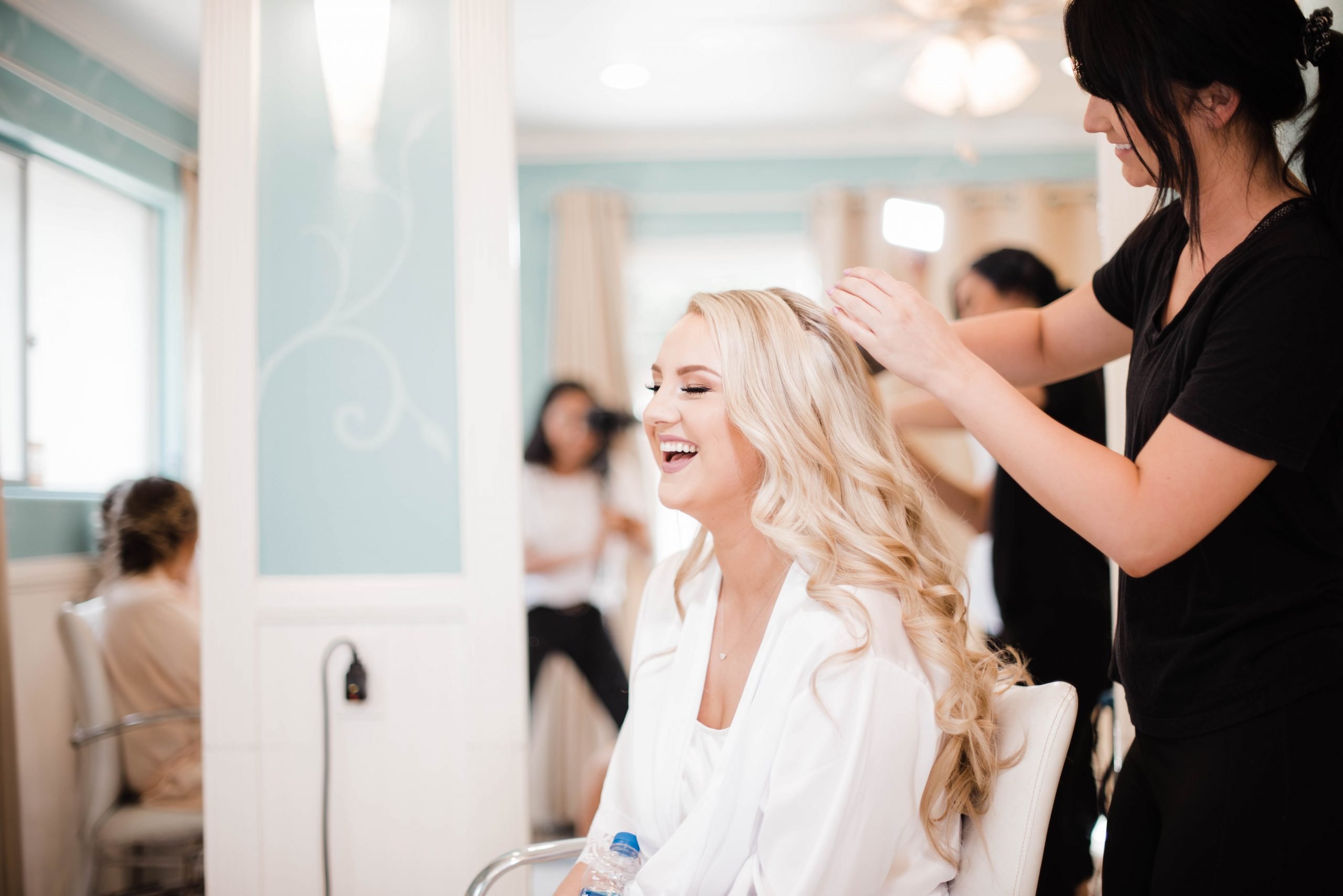 A hairstylist arranges the hair of a smiling woman seated in a salon chair, with other people visible in the background.