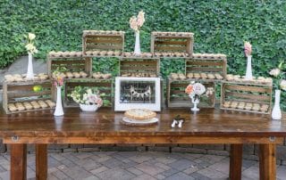 A wooden table displays pies and cookies in wooden crates, with white vases holding flowers, set against an ivy-covered wall outdoors.