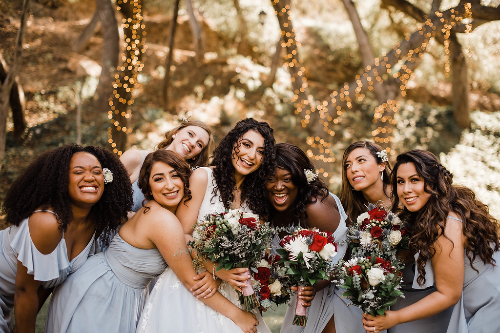 A bride and six bridesmaids in light blue dresses pose outdoors, smiling and holding bouquets with red and white flowers. Trees with string lights are in the background.