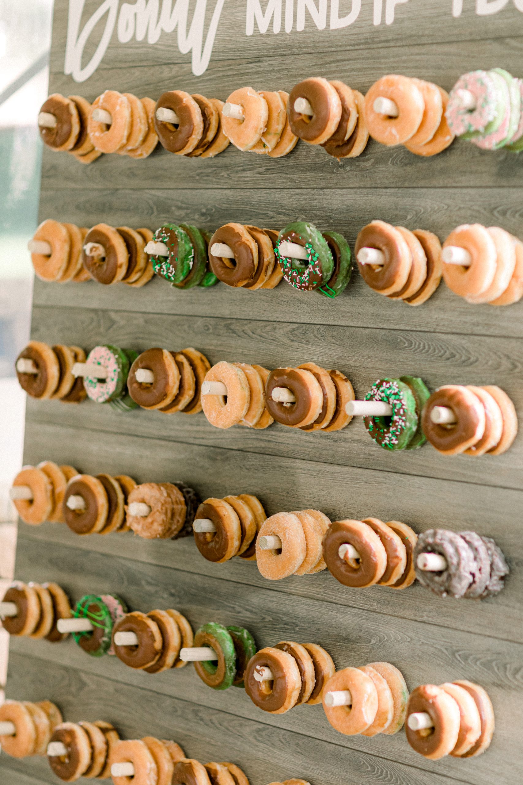 A wooden display board with rows of assorted donuts, some with chocolate or green icing and sprinkles, each stacked on individual pegs.