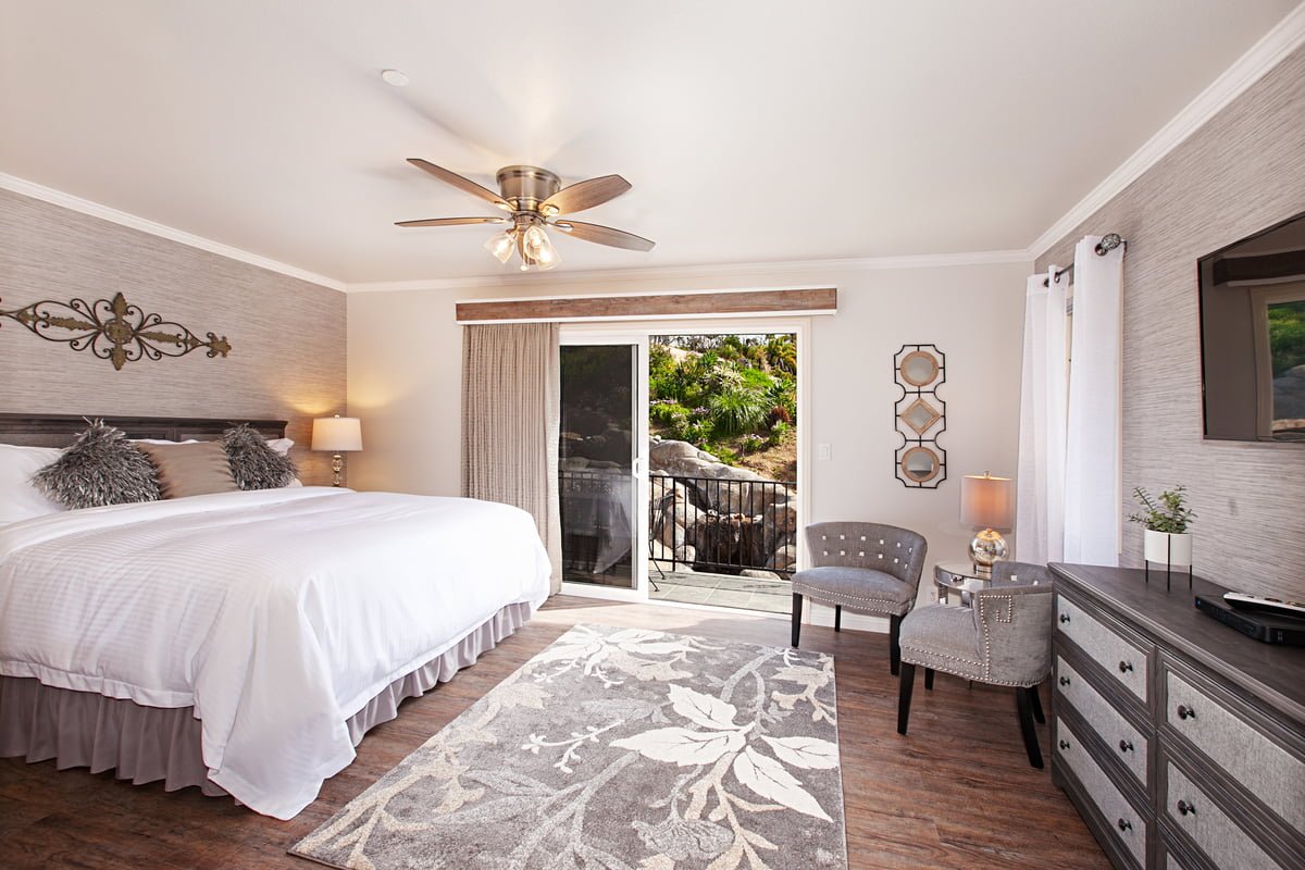 Modern bedroom with king bed, ceiling fan, dresser, two chairs, a rug, and sliding glass doors opening to an outdoor area with greenery.