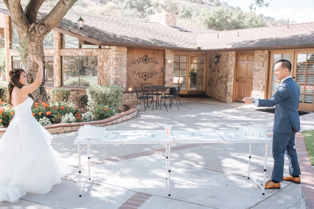 A bride and groom play a game of beer pong on a folding table outdoors in a courtyard, -1163 displayed nearby, with a house and garden in the background.