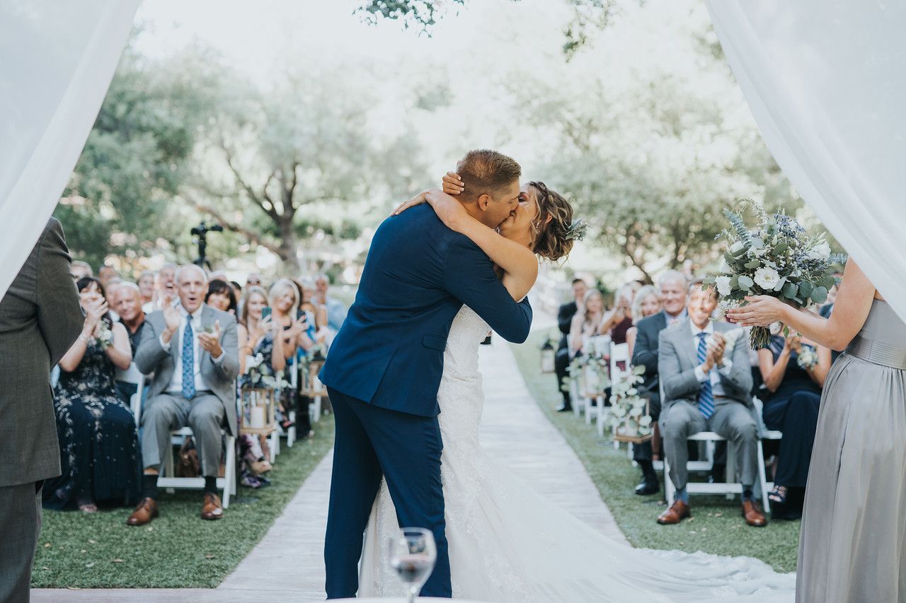 A bride and groom share a kiss at an outdoor wedding ceremony, while guests seated on either side applaud and celebrate the -1163 moment.