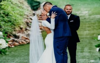 A bride and groom kiss under a chandelier during their outdoor wedding ceremony, while an officiant stands behind them and smiles.