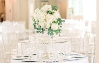 A round table with a tall floral centerpiece featuring white flowers and greenery, set for a formal event in a bright, elegant room with white chairs and décor.