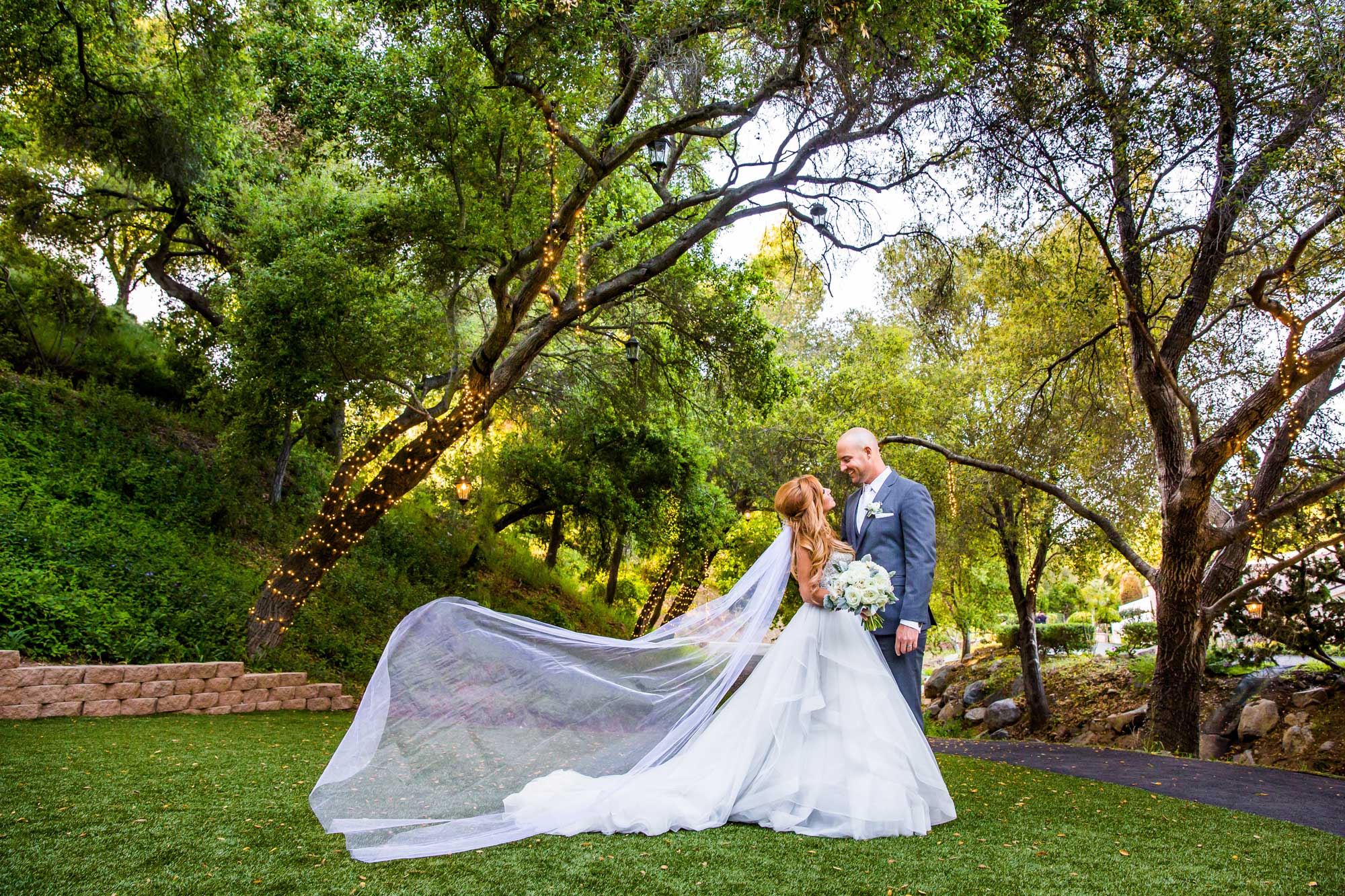 A bride and groom stand outdoors on grass, surrounded by trees and soft -1163 lighting, with the bride’s veil flowing behind her and strings of lights wrapped around a tree trunk.