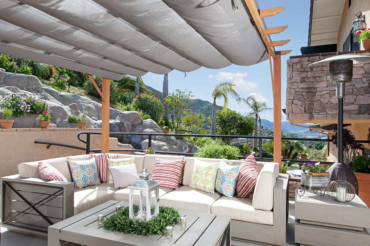 Outdoor patio -1163 features a cushioned sectional sofa, striped and patterned pillows, coffee table with decorative lantern, potted plants, sunshade, and a scenic view of hills in the background.