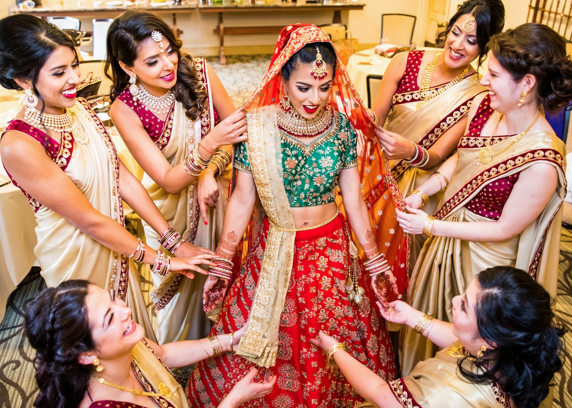 A bride in a green and red traditional outfit stands surrounded by six bridesmaids in cream and red sarees, all smiling and adjusting her attire.