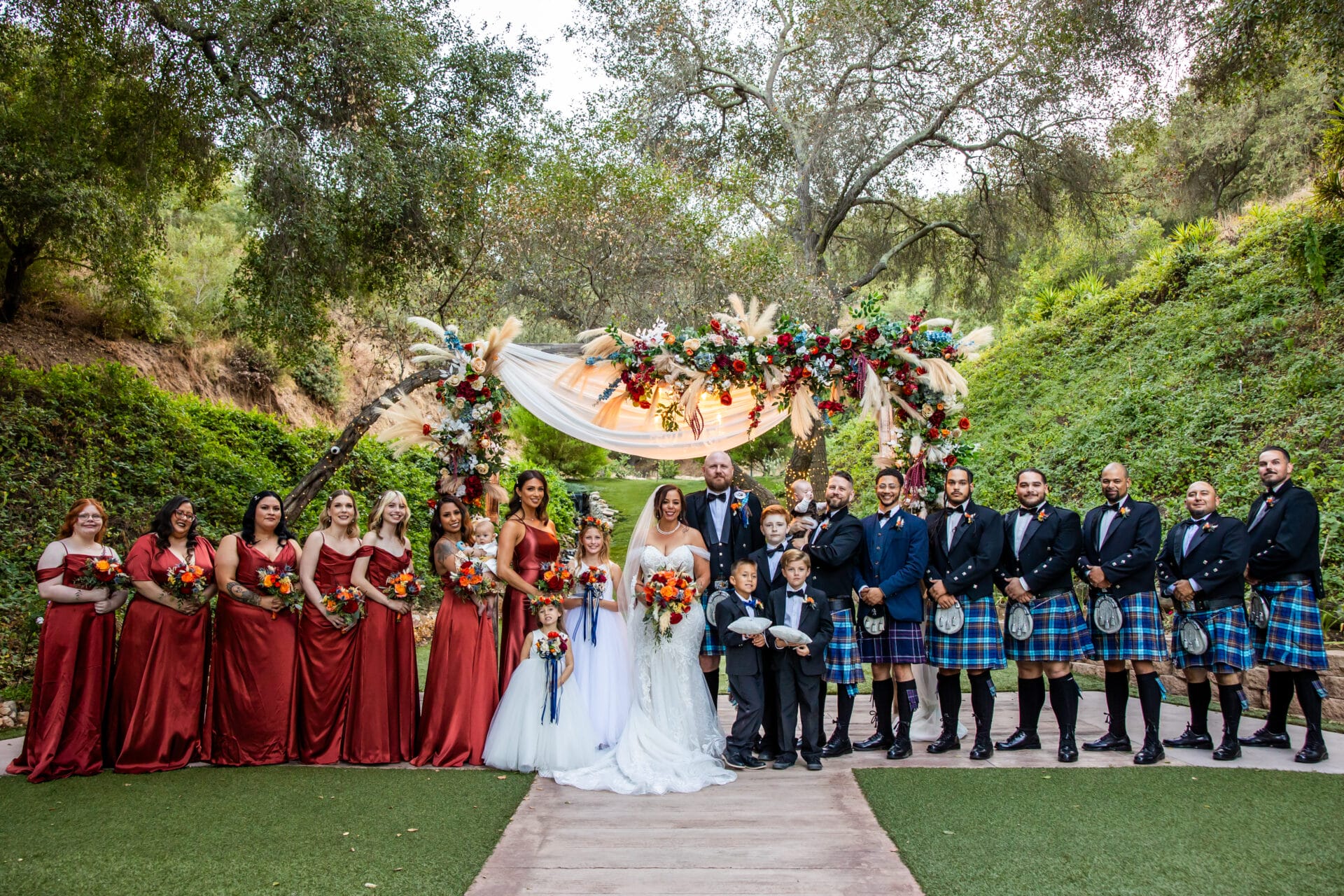 A wedding party poses outdoors; bridesmaids wear red dresses, groomsmen wear kilts, and the bride and groom stand under a floral arch with trees in the background.
