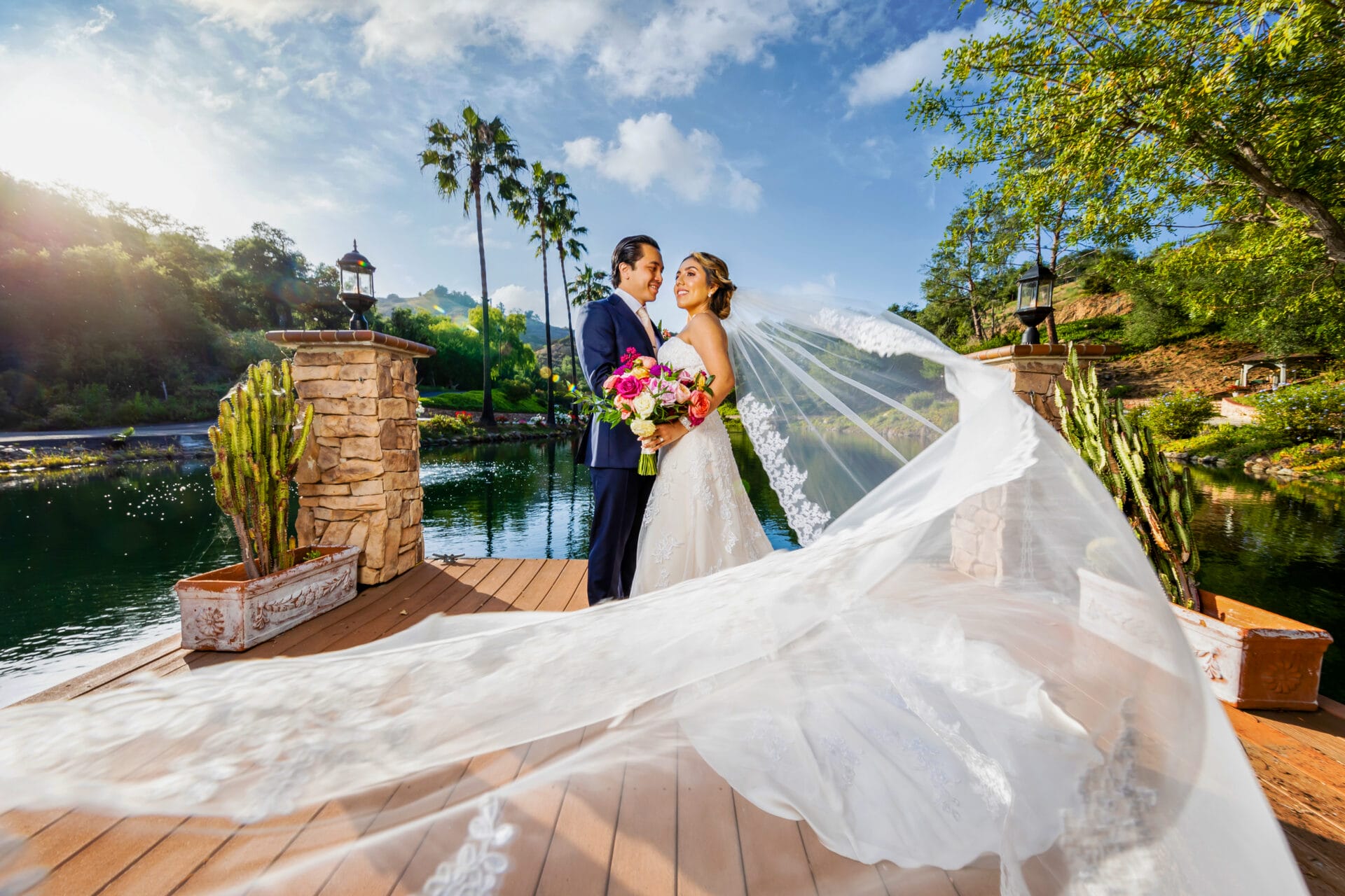 A bride and groom stand on a wooden dock by a pond, holding a bouquet, with the bride's veil flowing dramatically in the breeze.